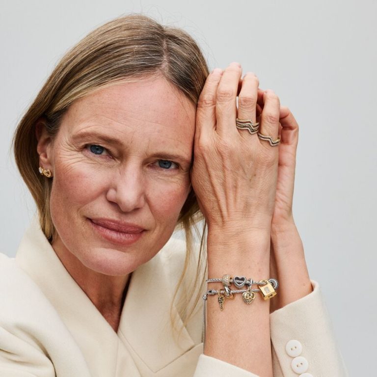 Person wearing layered gold and silver charm bracelets and rings, shown in a close-up portrait.